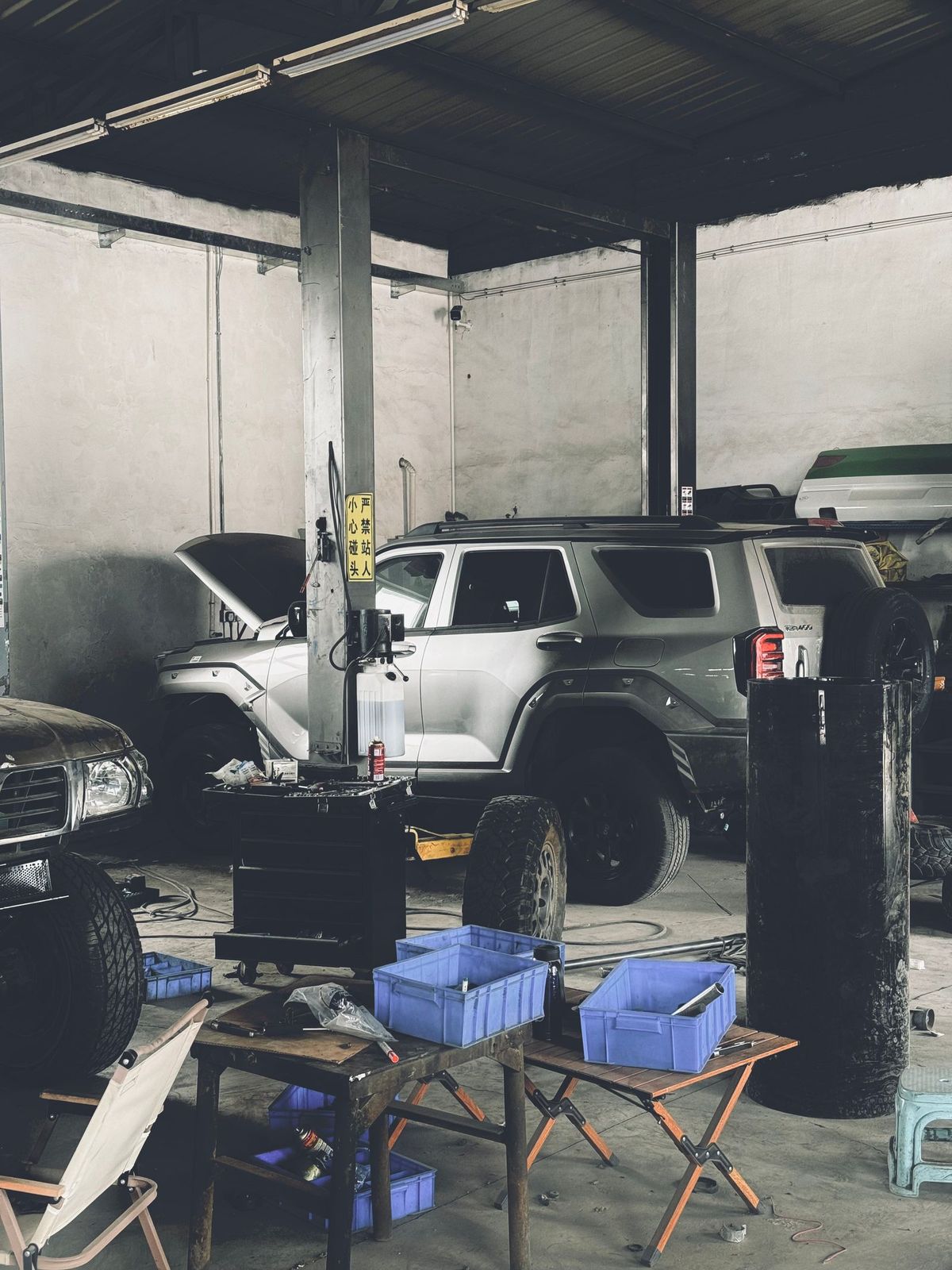 A workshop with a car's bonnet open beside a lift column, tools and parts crates around it
